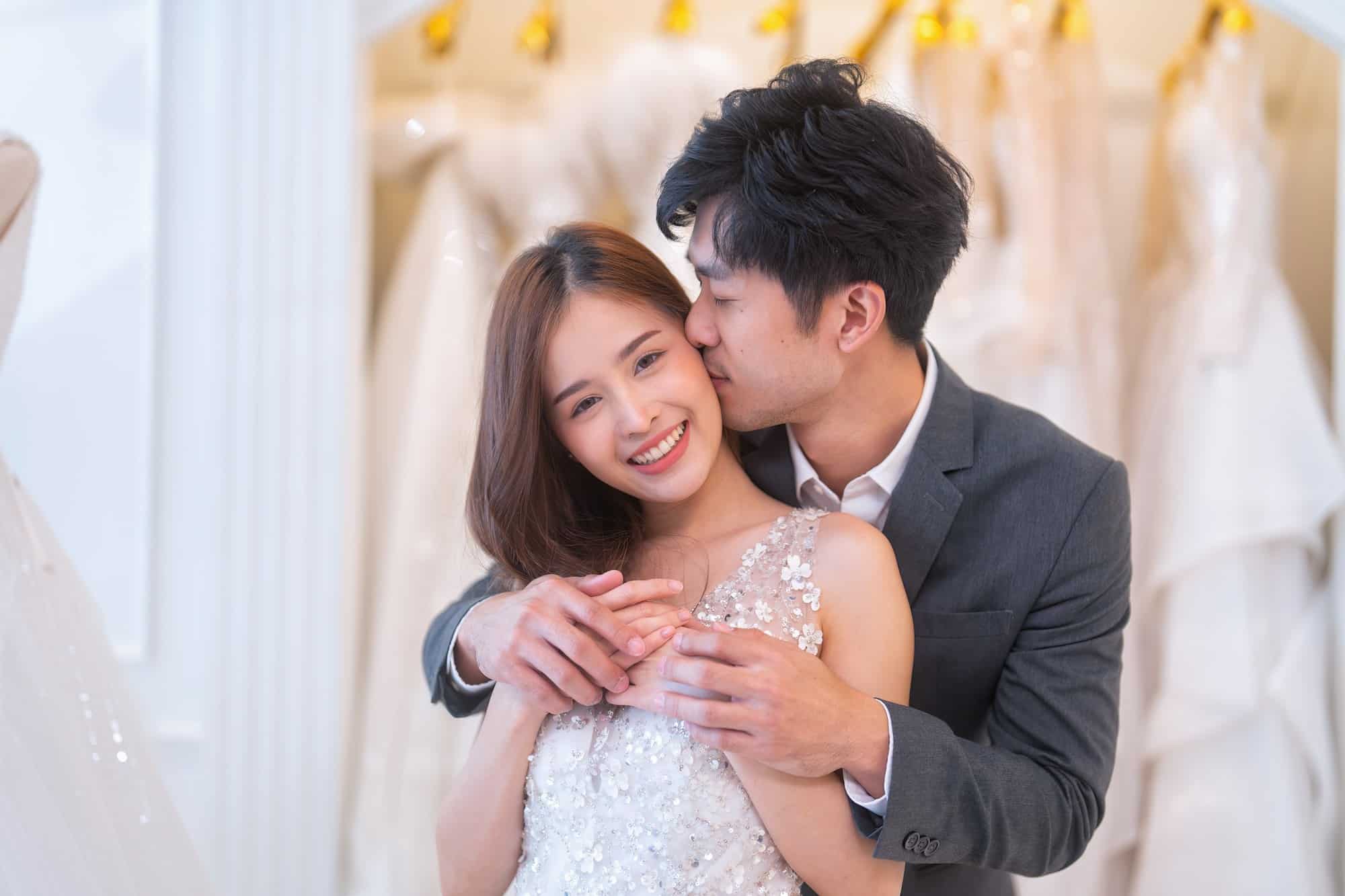 Beautiful young brunette love couple choosing wedding dress in a bridal salon a man stand hugging