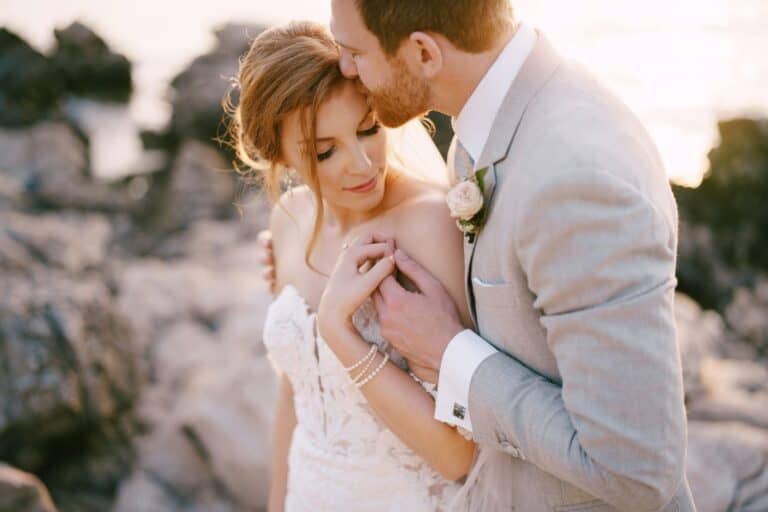 Groom kisses bride, hugging her shoulders on the rocky seashore